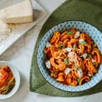 creamy gochujang pasta in blue patterned bowl on leaf shaped placemat beside dish with kimchi and dish with shredded toscano cheese