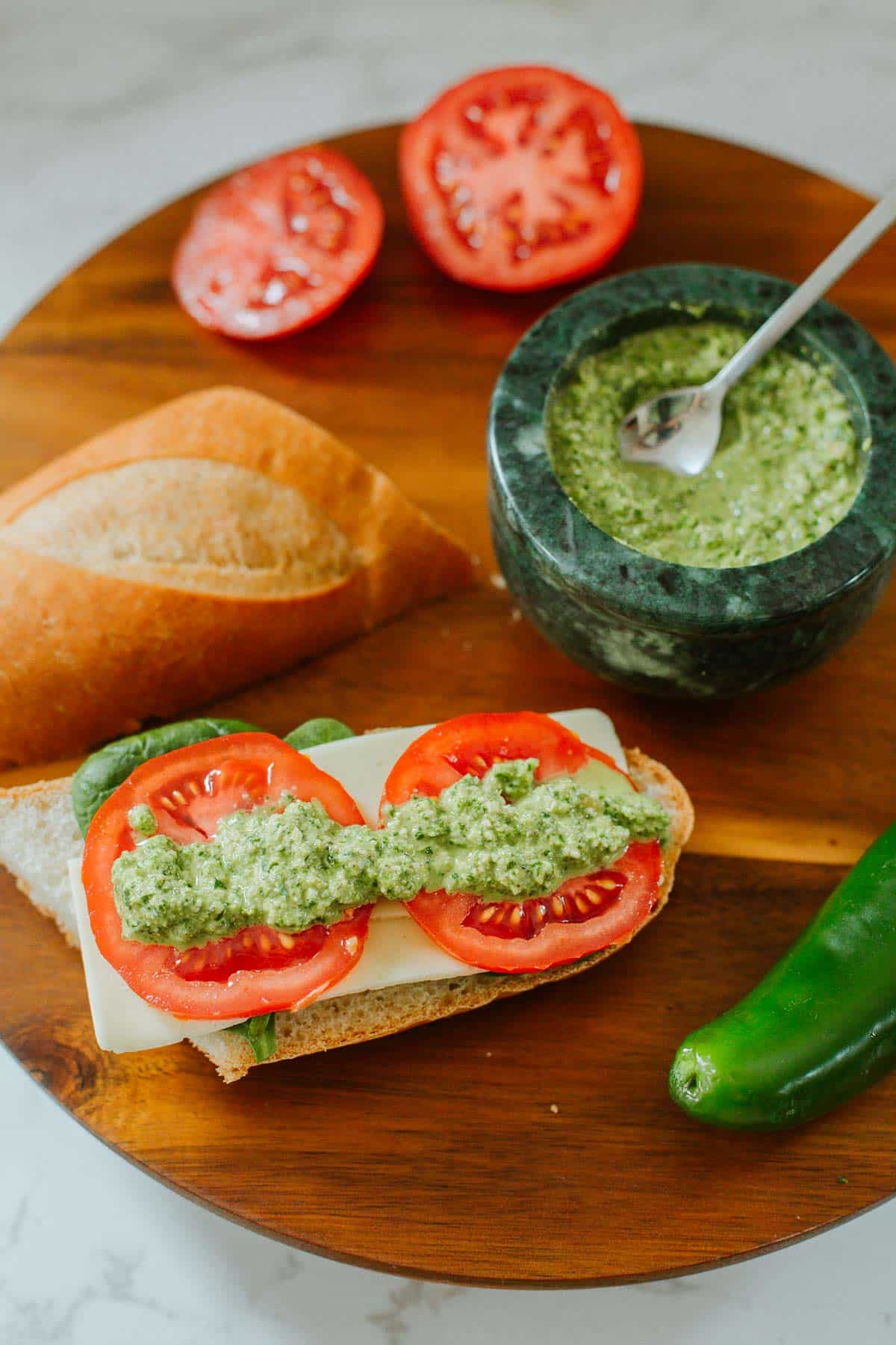 spicy jalapeno pesto over a tomato sandwich beside baguette half, bowl of pesto, and sliced tomatoes