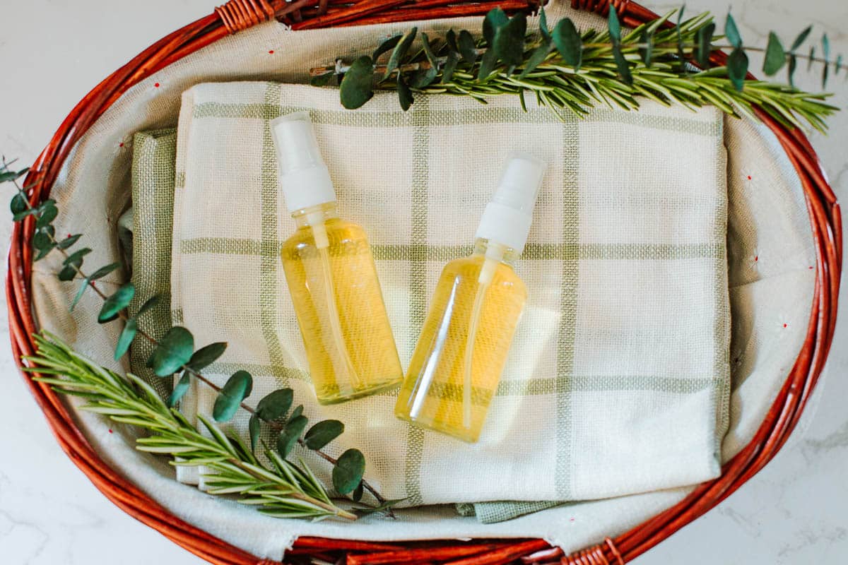 two spray bottles with rosemary water for hair in a basket alongside fresh rosemary and eucalyptus sprigs.