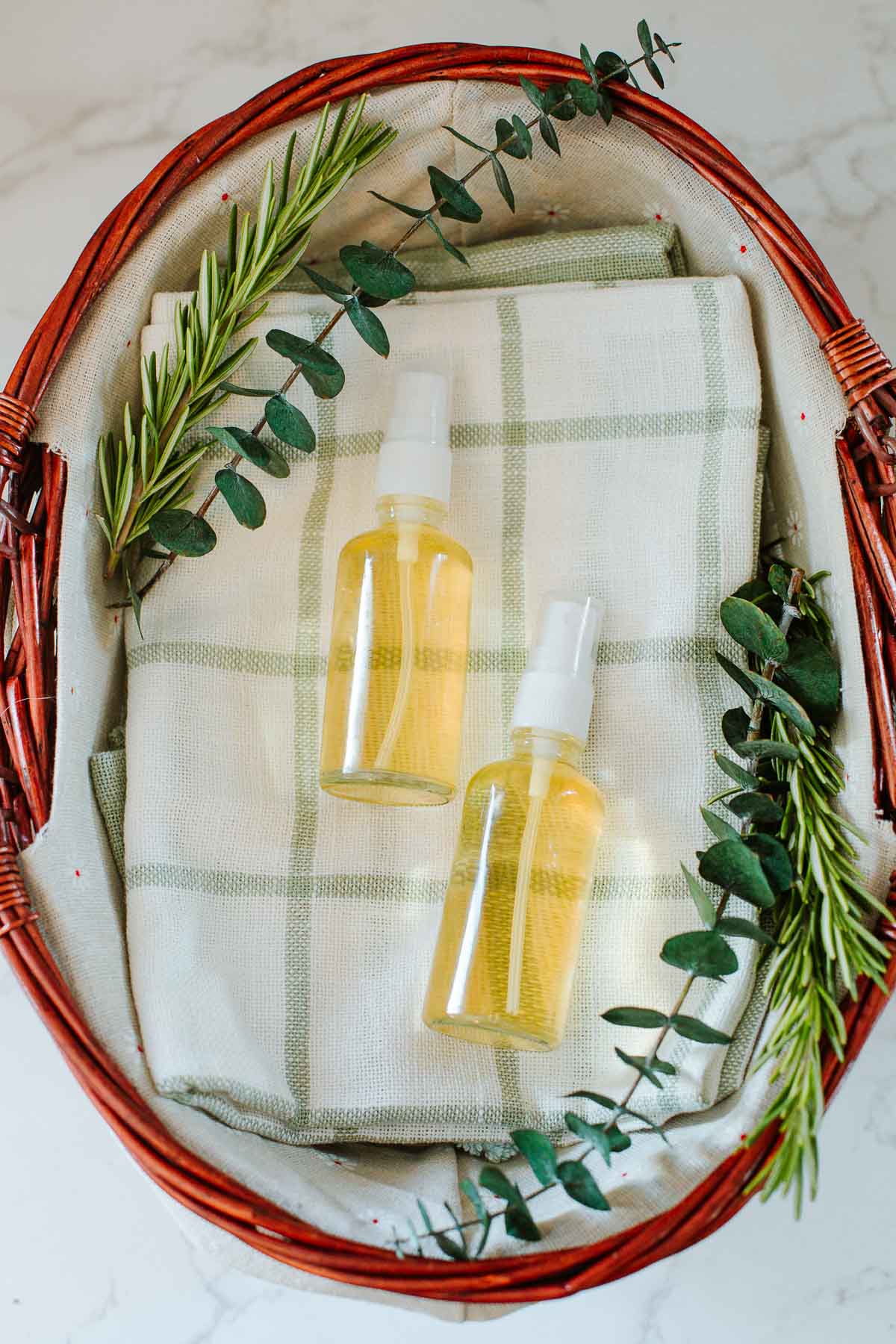two spray bottles with rosemary water for hair in a basket alongside fresh rosemary and eucalyptus sprigs