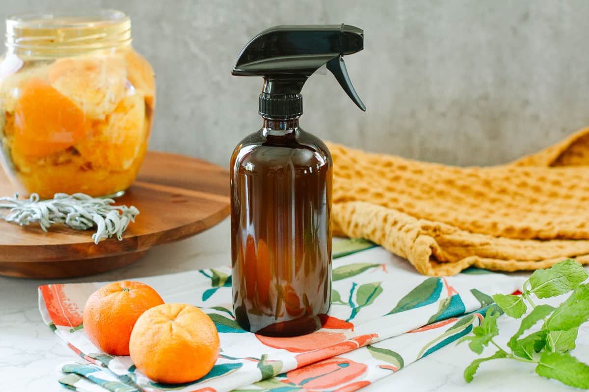 orange peel & vinegar cleaner in spray bottle beside colorful towels, fruits, herbs, jar of peels