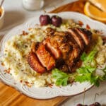 sliced air fryer duck breast with cherry sauce on a bed of mashed potatoes, surrounded by fresh cherries, orange slices, herbs, bowl of sauce.