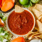 guajillo salsa in bowl with chip alongside more chips, veggies, wooden spoon.