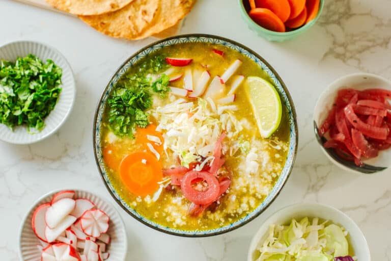 loaded pork pozole verde surrounded by cilantro, radish, onion, cabbage, carrots, tortillas.