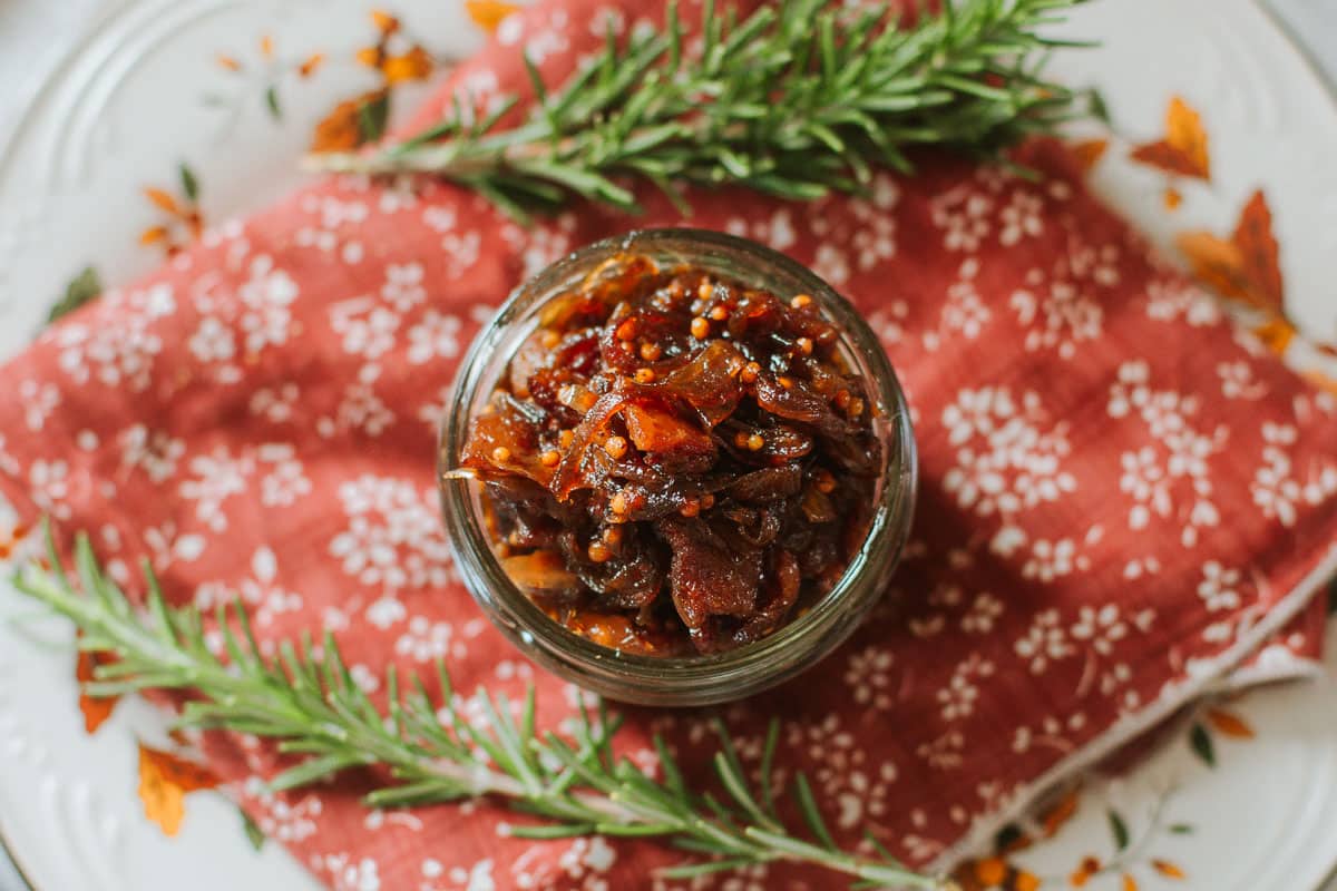 caramelized onion jam in mason jar on floral placemat with fresh rosemary sprigs.