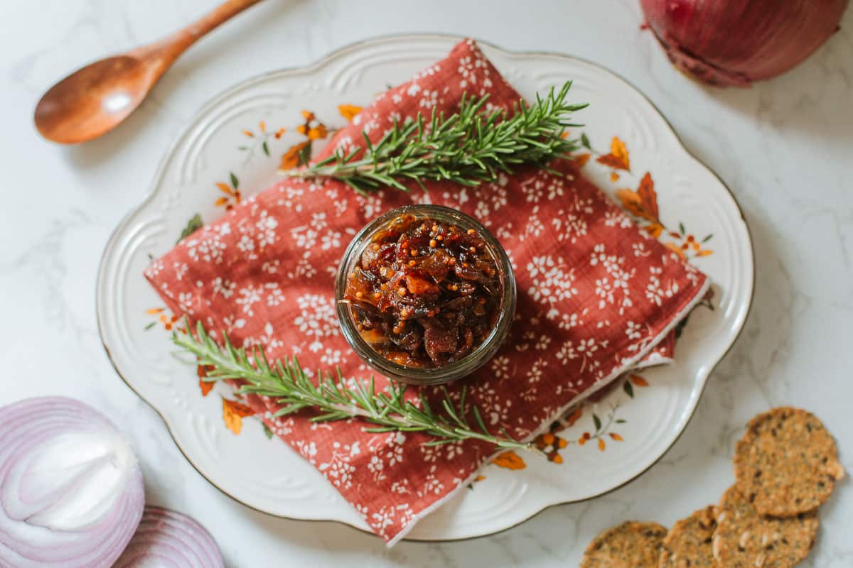 caramelized onion jam in mason jar on floral placemat with fresh rosemary sprigs, spoon, red onion slices, crackers.