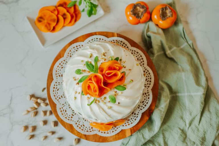 persimmon cake topped with persimmon slices formed into "roses" beside fresh fuyu persimmons, pistachios, and tea towel.