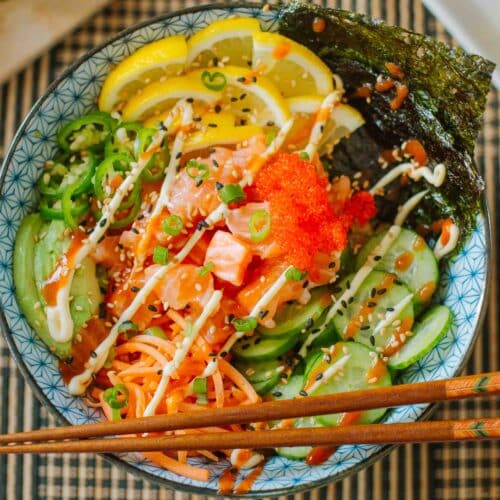 salmon sushi bowl loaded with fresh veggies, lemon slices, seaweed, etc. with chopsticks resting on the edge.