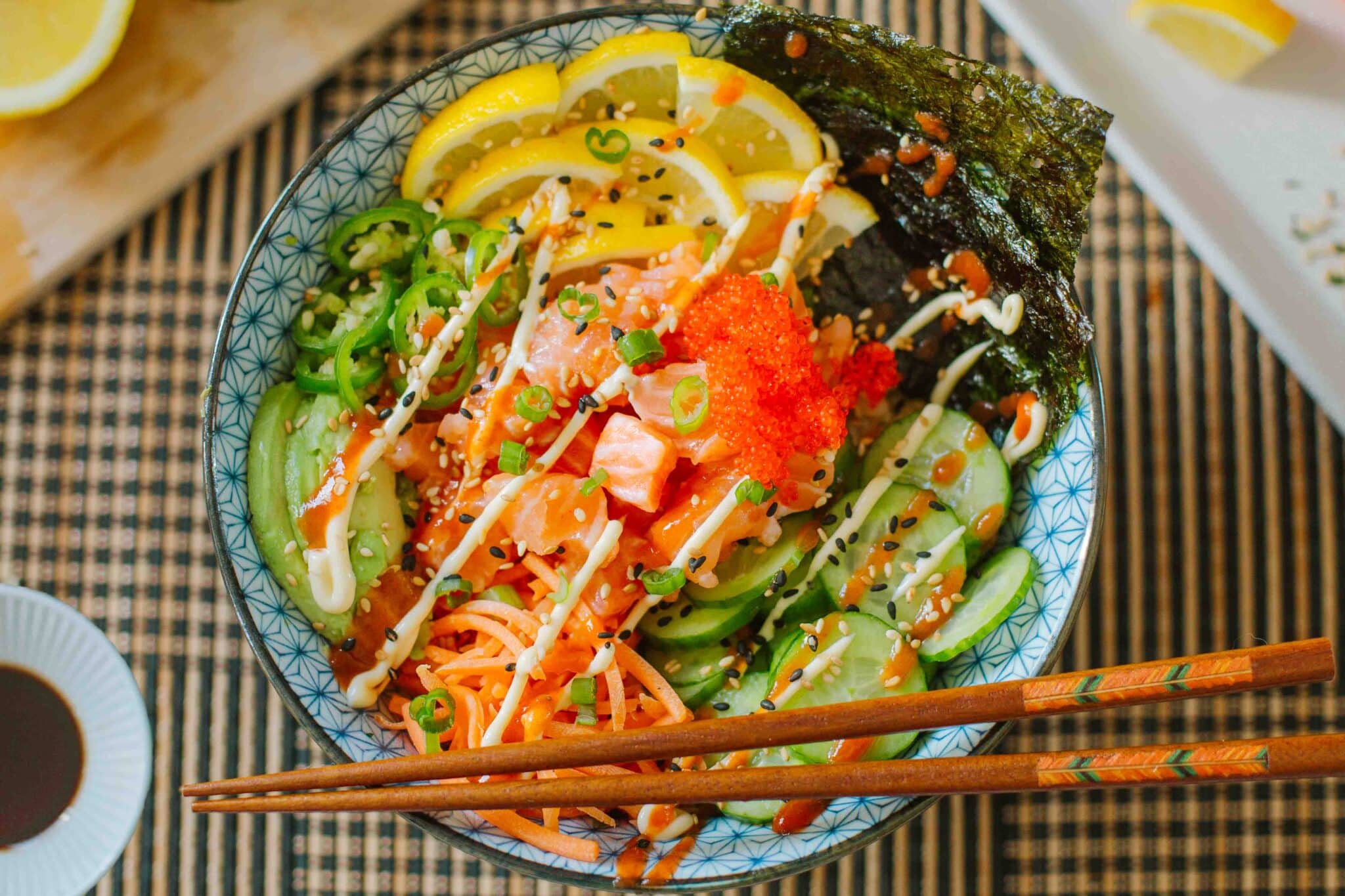 salmon sushi bowl loaded with fresh veggies, lemon slices, seaweed, etc. with chopsticks resting on the edge.