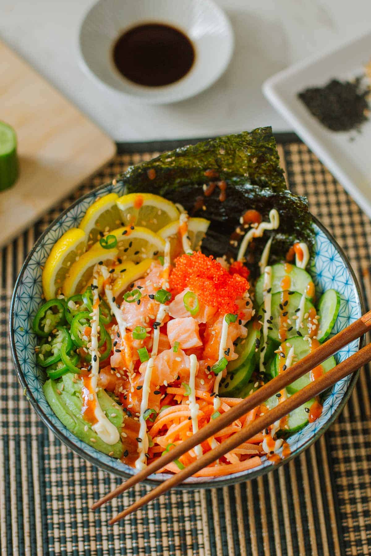 salmon sushi bowl loaded with fresh veggies, lemon slices, seaweed, etc. with chopsticks resting on the edge.