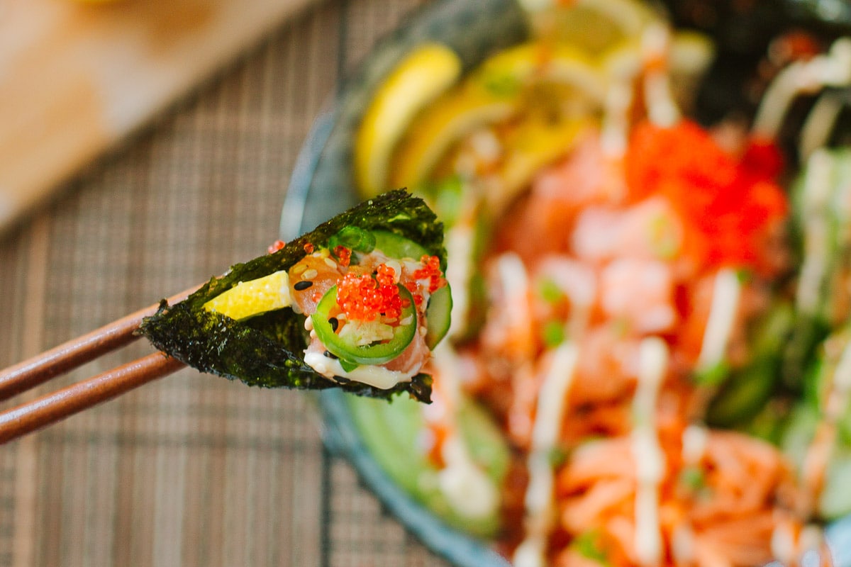 nori sheet full of salmon, masago, and veggies being held between chopsticks above sushi bowl.