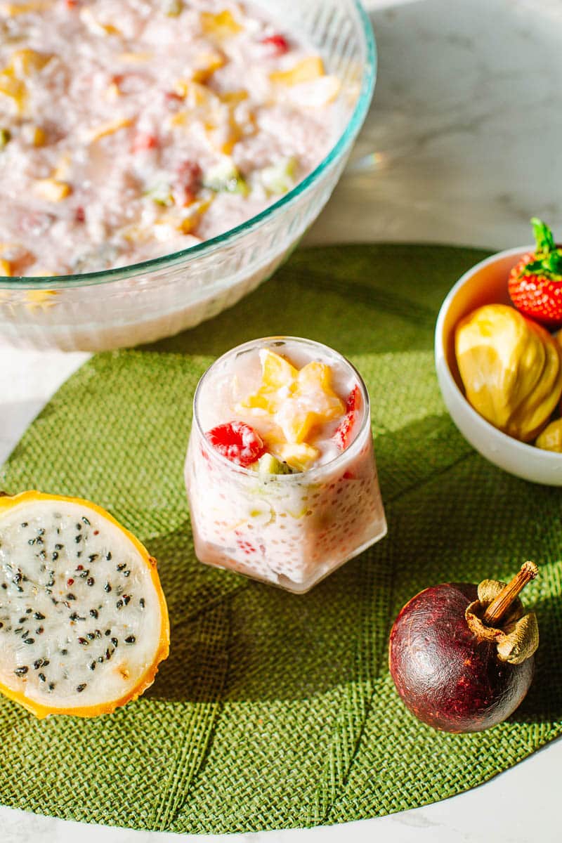 coconut milk sago in cup beside bowl and surrounded by fresh tropical fruits.